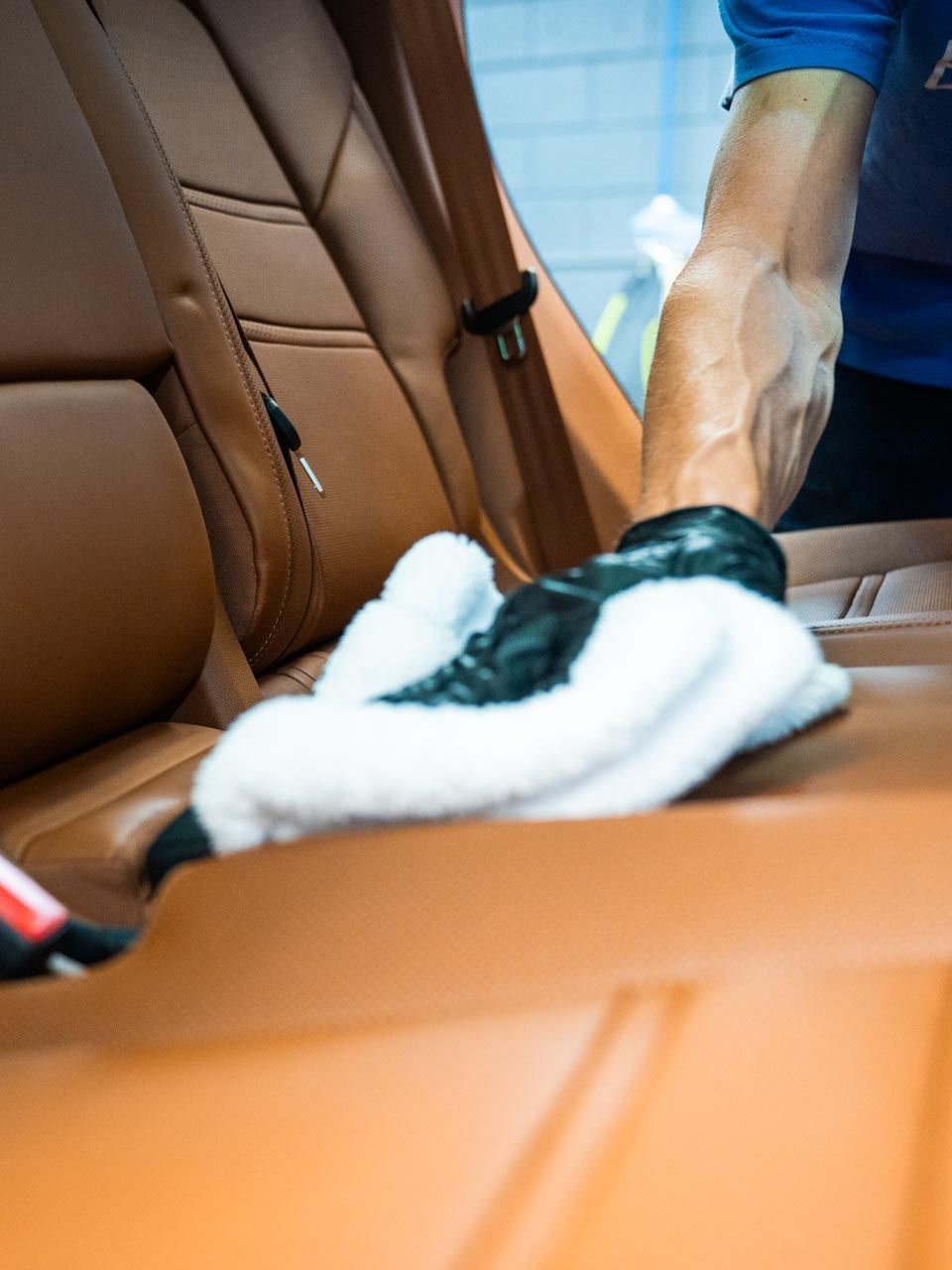 A person's hand wearing a black glove wipes down a tan car seat with a white cloth.