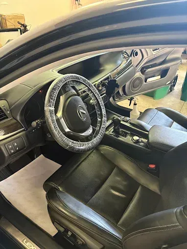 Interior of a black Lexus car, driver's side. Steering wheel covered in plastic. Black leather seats.