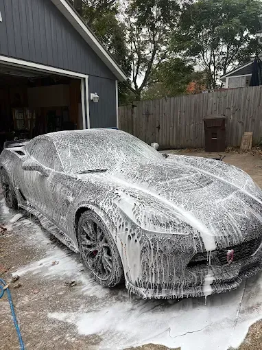Gray sports car covered in white soap suds being washed in front of a garage.