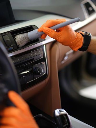 Person wearing orange gloves dusting a car's air vents with a gray brush inside the vehicle.