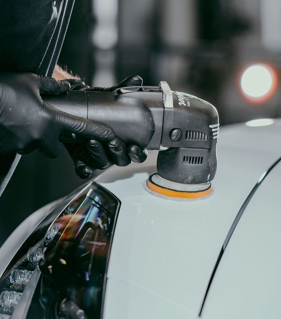 A person wearing black gloves polishes a white car with an electric polisher.