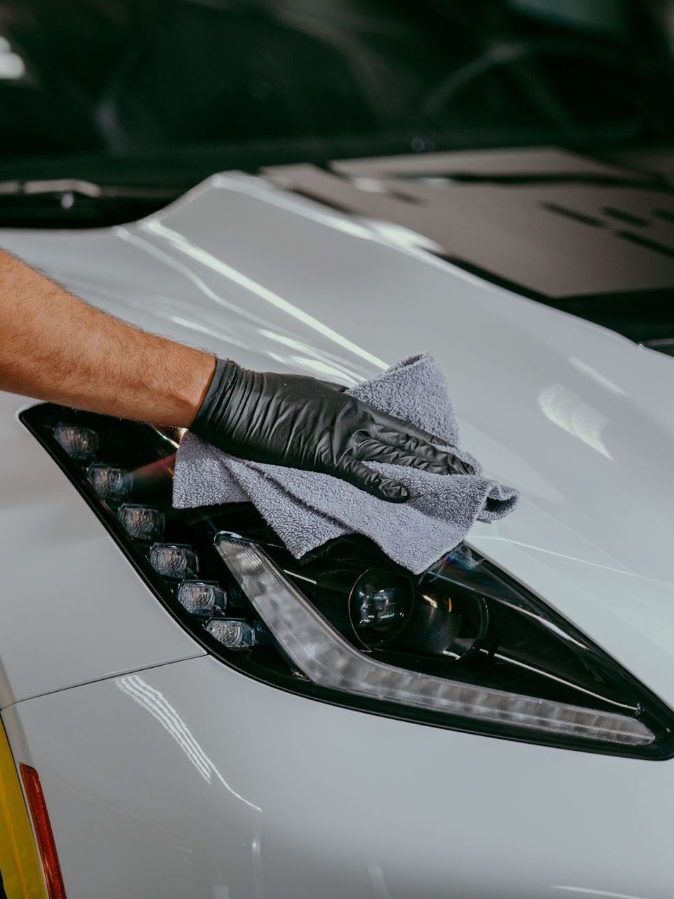 A person wearing a black glove wipes a white car headlight with a gray microfiber cloth.