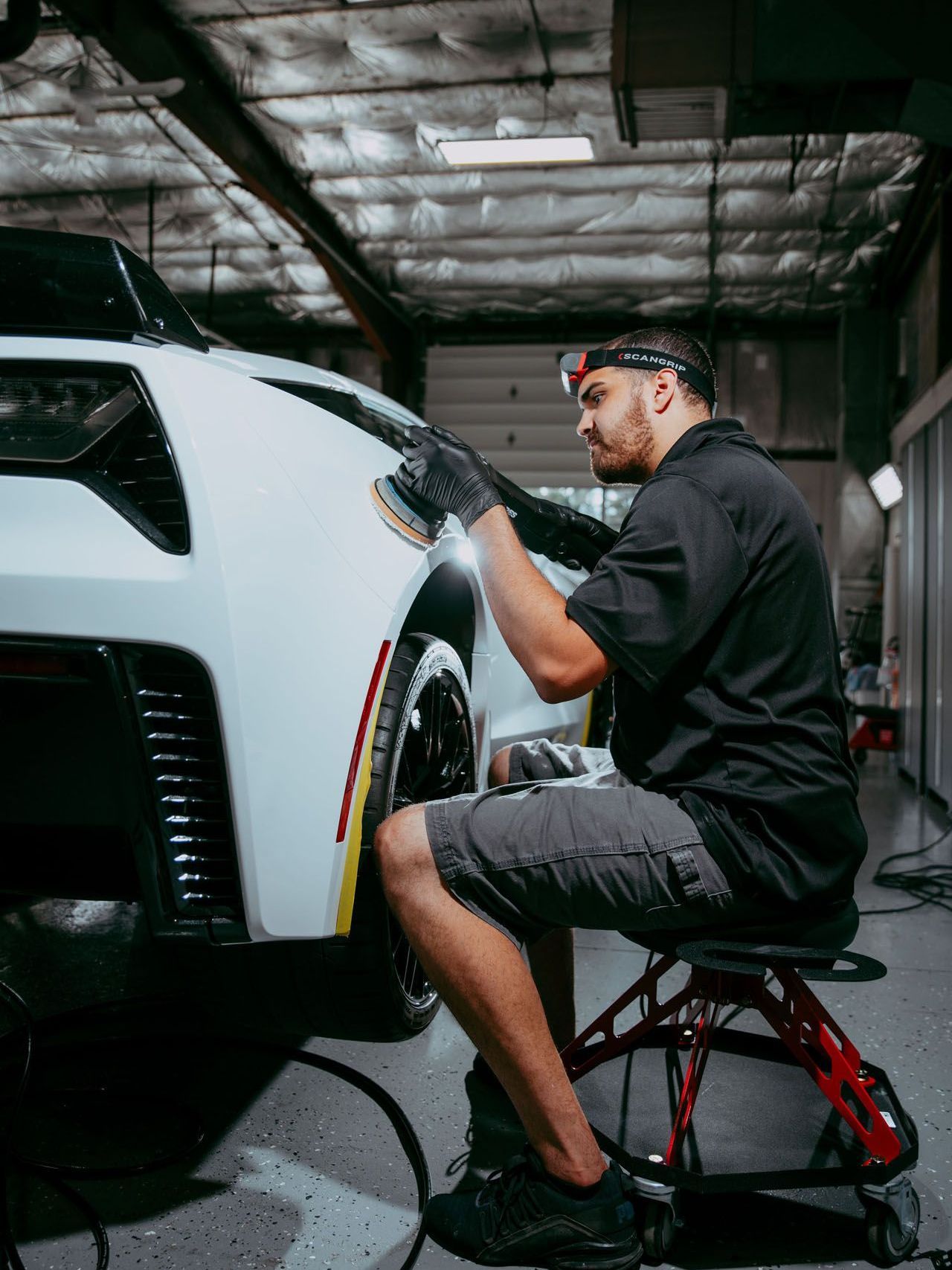 Man polishing a white car in a garage. He wears a headlamp, gloves, and sits on a stool.