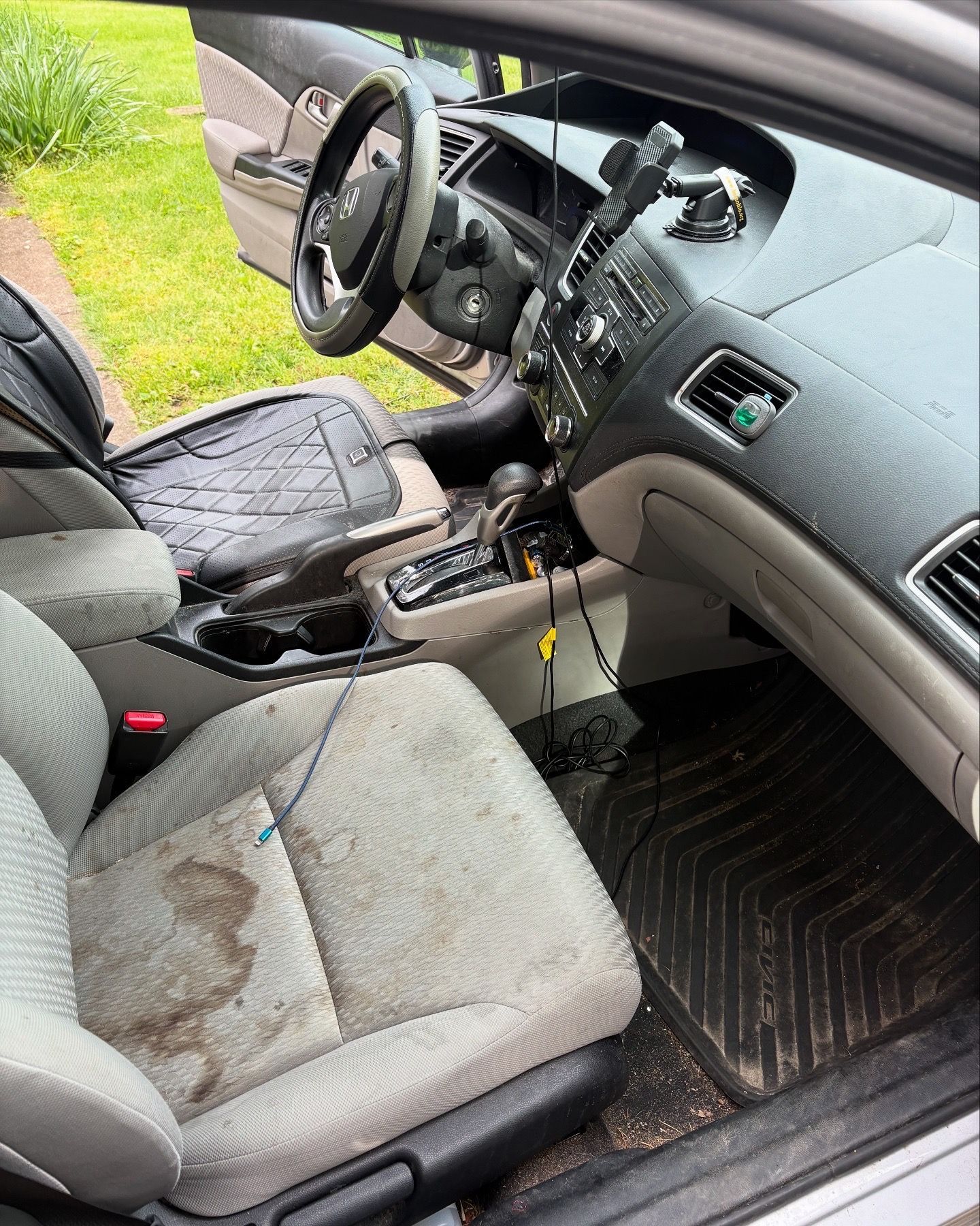 Dirty car interior: stained front seat, open driver's side door, black floor mats, steering wheel visible.