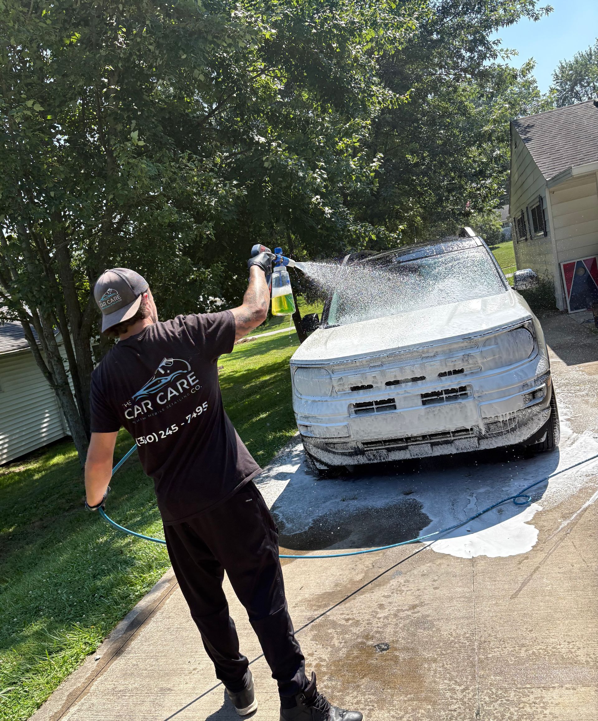 Woman with pink hair washes a dark car with a pressure washer in a garage. Water sprays across the car.