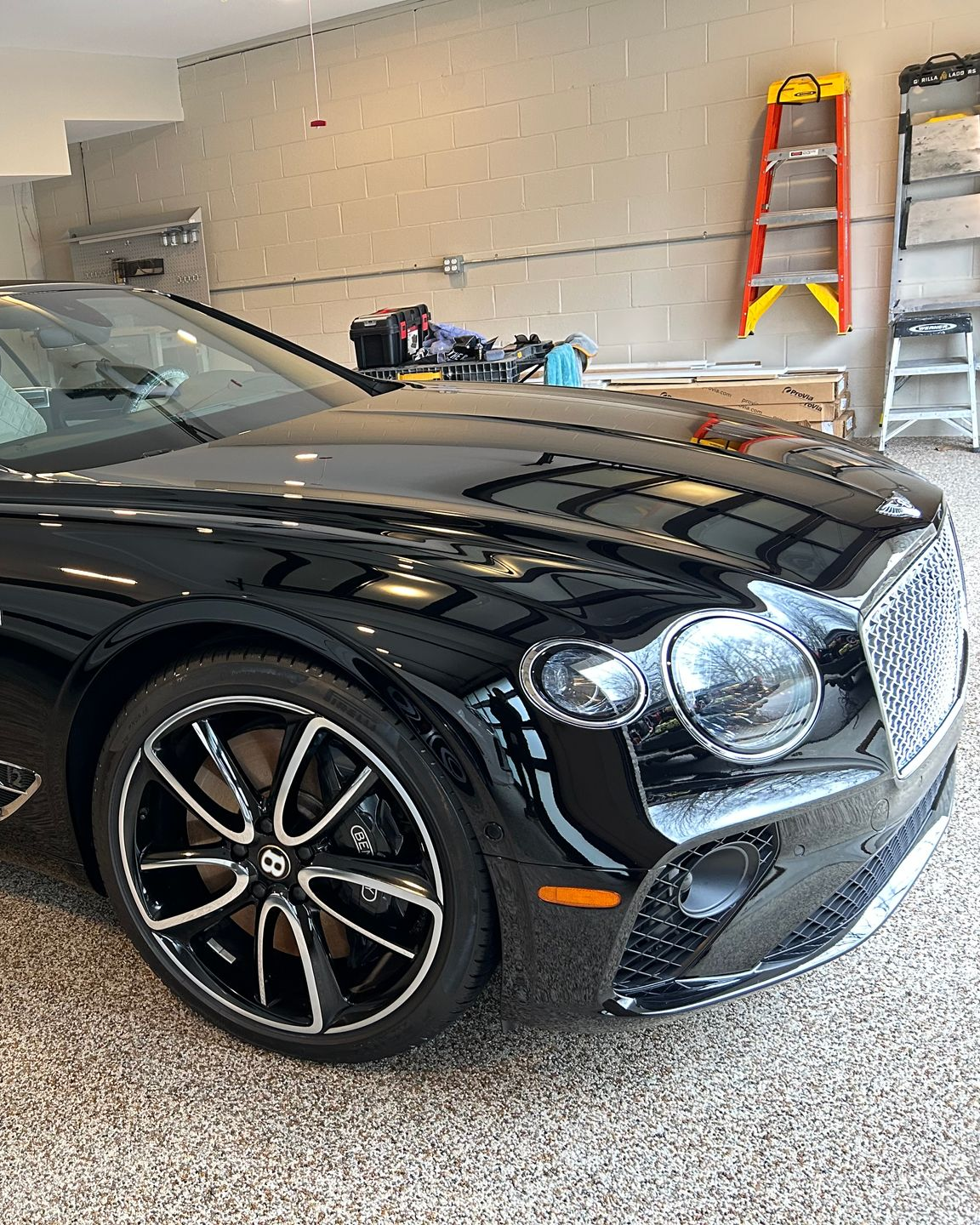 A glossy black luxury Bentley parked in a garage with a speckled floor, featuring prominent alloy wheels and headlights.