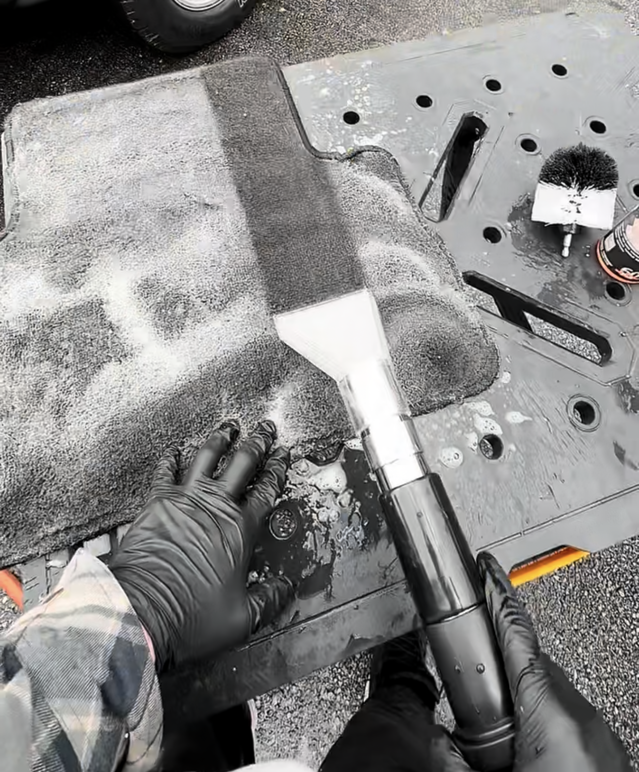 Person vacuuming the brown carpet of a car interior. The person wears black gloves and holds an orange hose.
