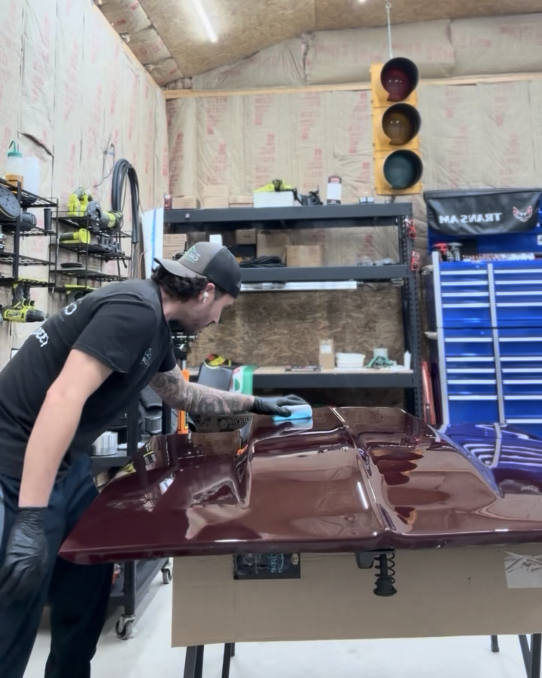 Man polishing a maroon car hood in a workshop; traffic light overhead.
