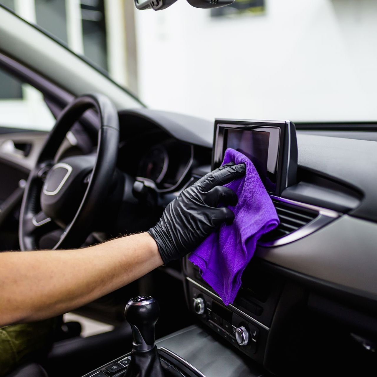 A person wearing a black glove wipes a car's dashboard screen with a purple cloth.