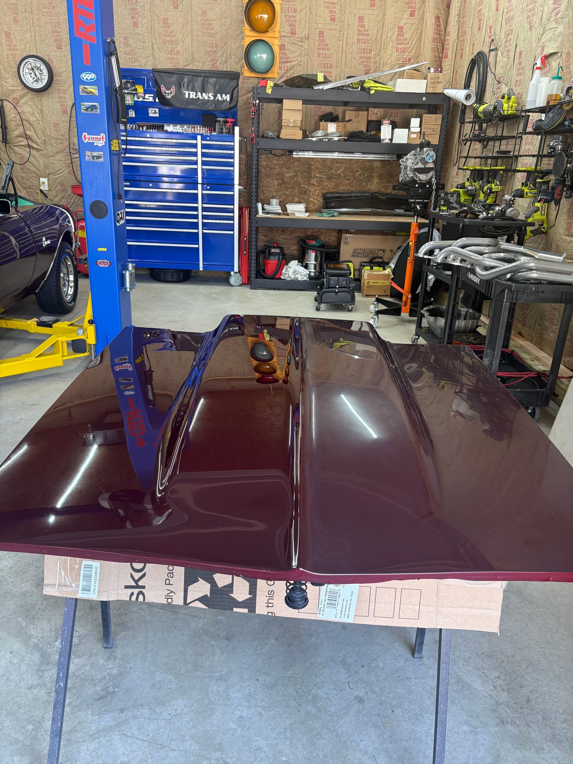 Maroon car hood on a stand in a garage. Workshop with tools and car lift in background.