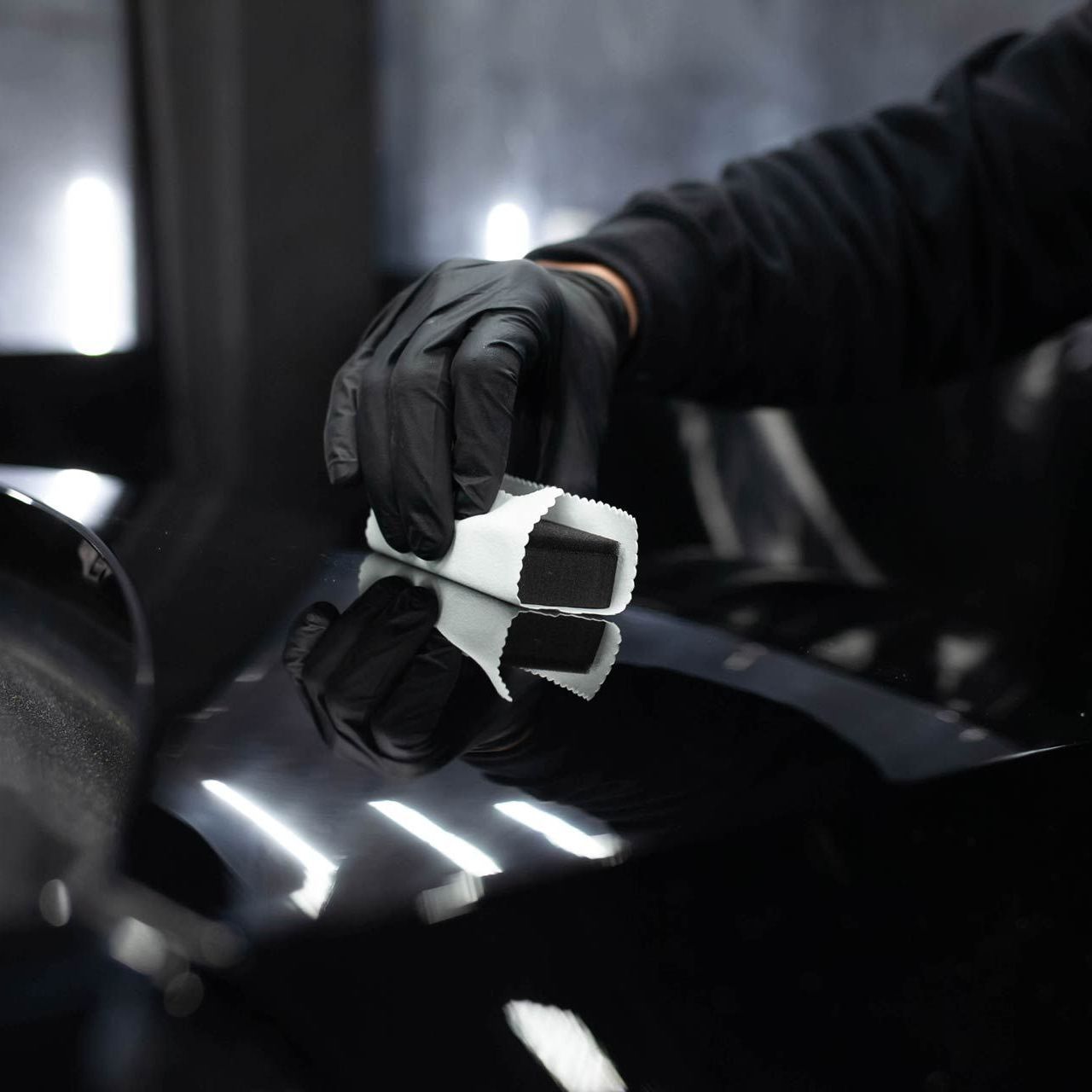 A person wearing black gloves applies a ceramic coating to a shiny black surface, likely a car.