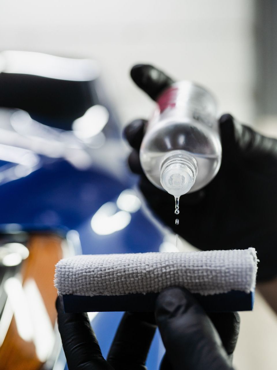 Gloved hands applying liquid from a bottle onto a microfiber applicator pad in front of a blue car.