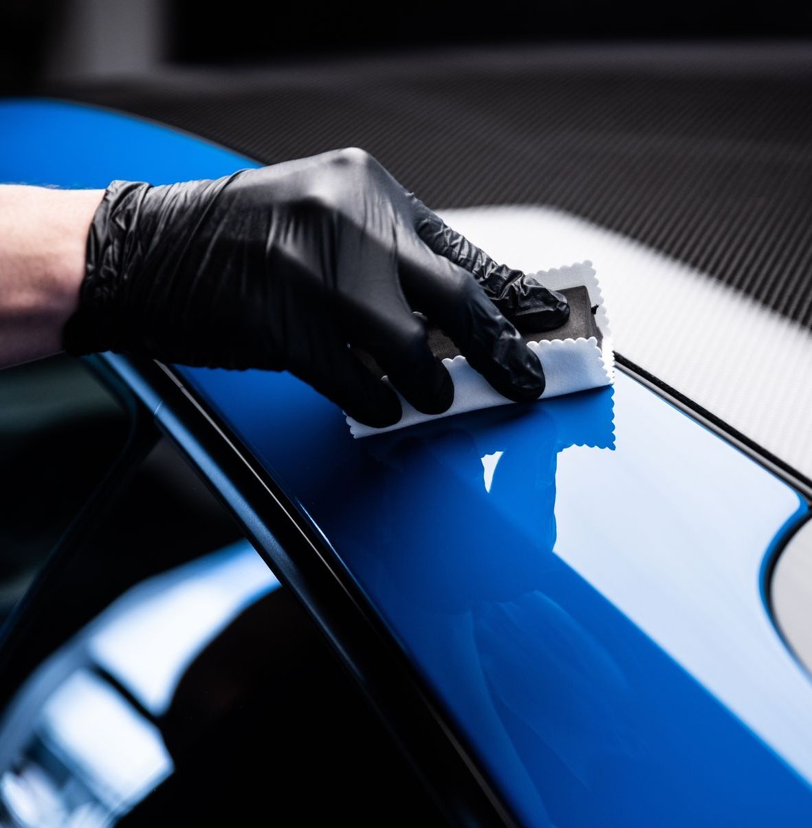 Gloved hand applying ceramic coating to a shiny blue car surface with a white applicator.
