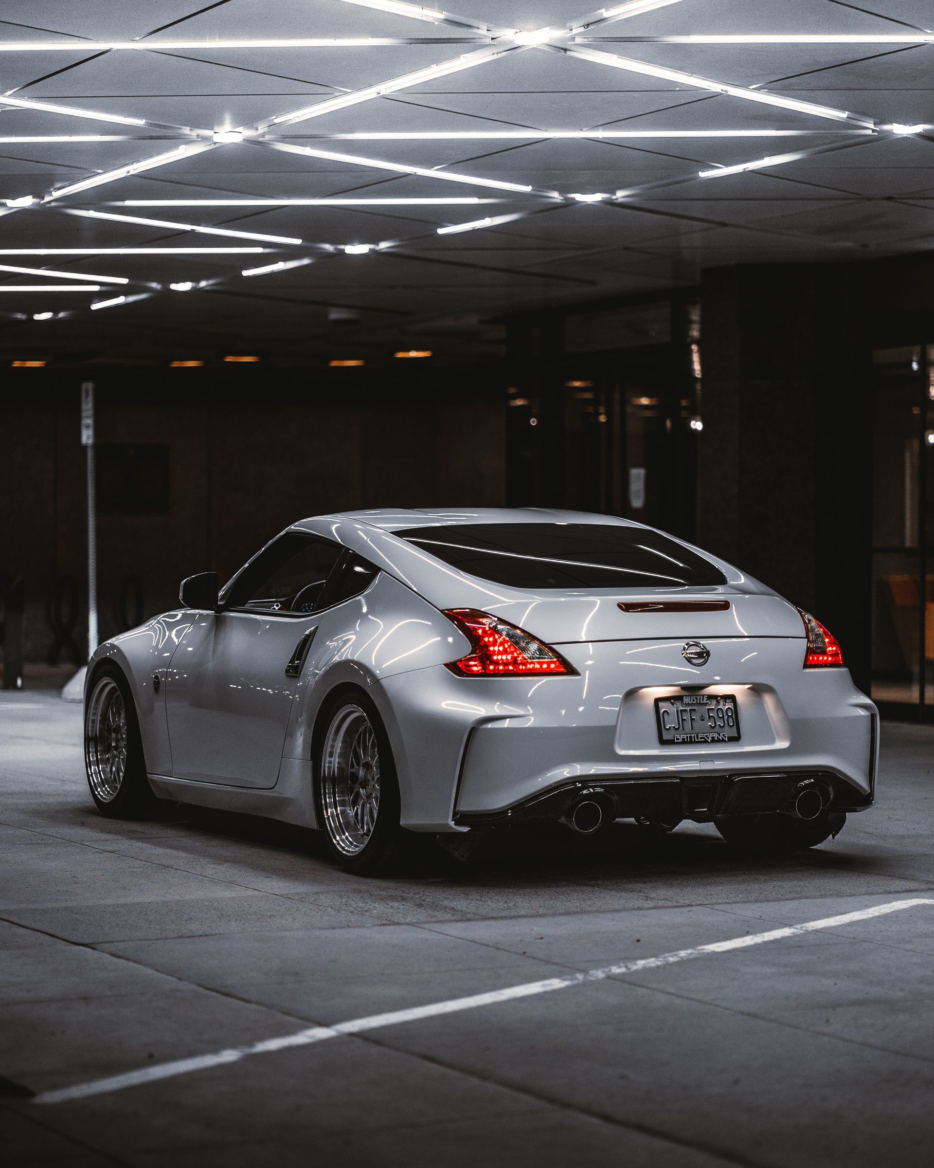 Silver Nissan 370Z sports car parked indoors under geometric ceiling lights. The car's rear is facing the viewer.