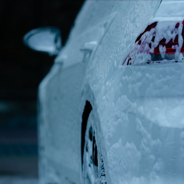 White car covered in thick, white foam, partially obscuring the vehicle's shape. Rear tire and taillight visible.