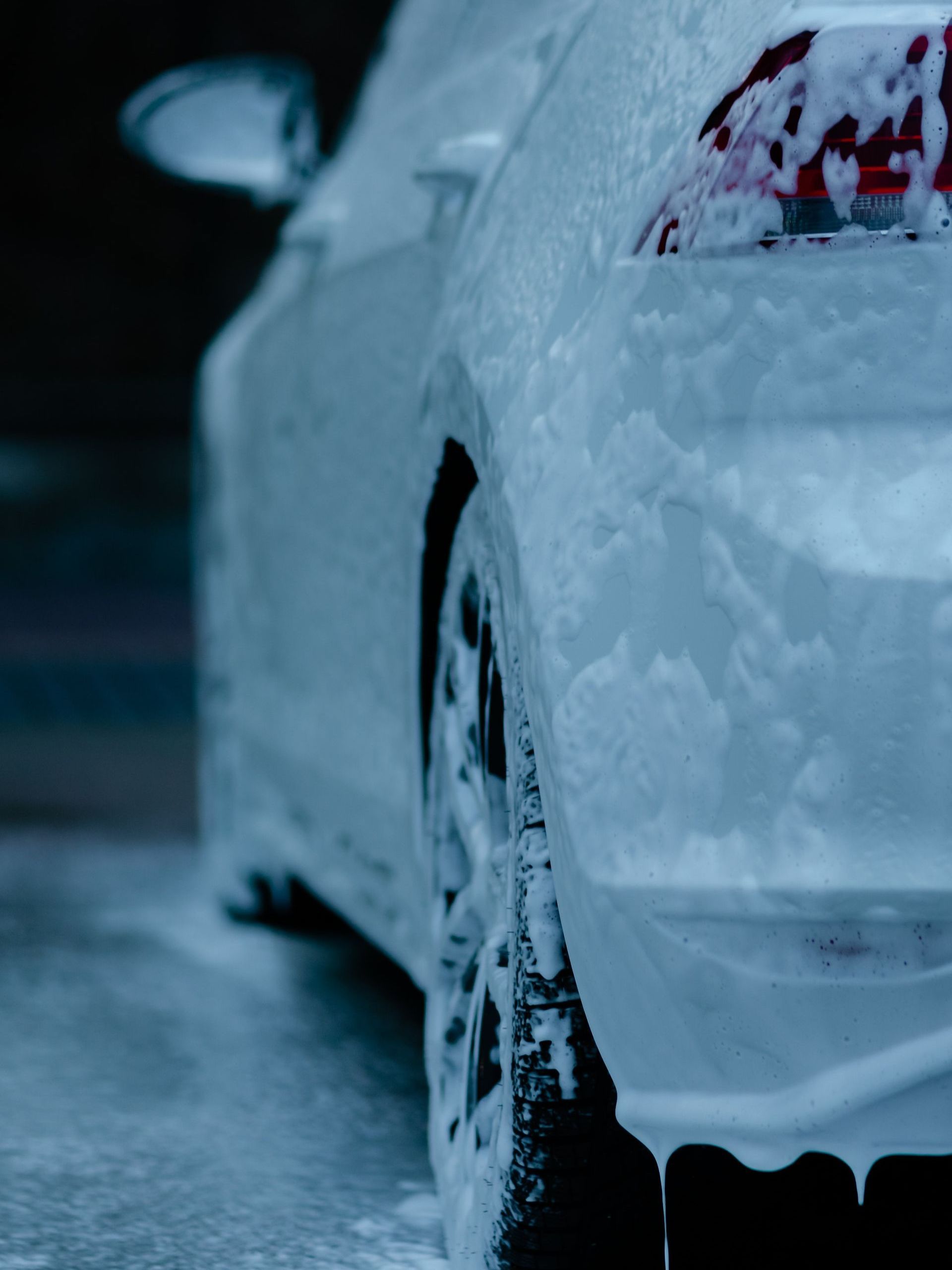 White car covered in thick, white foam at a car wash. Tail light and tire are visible.