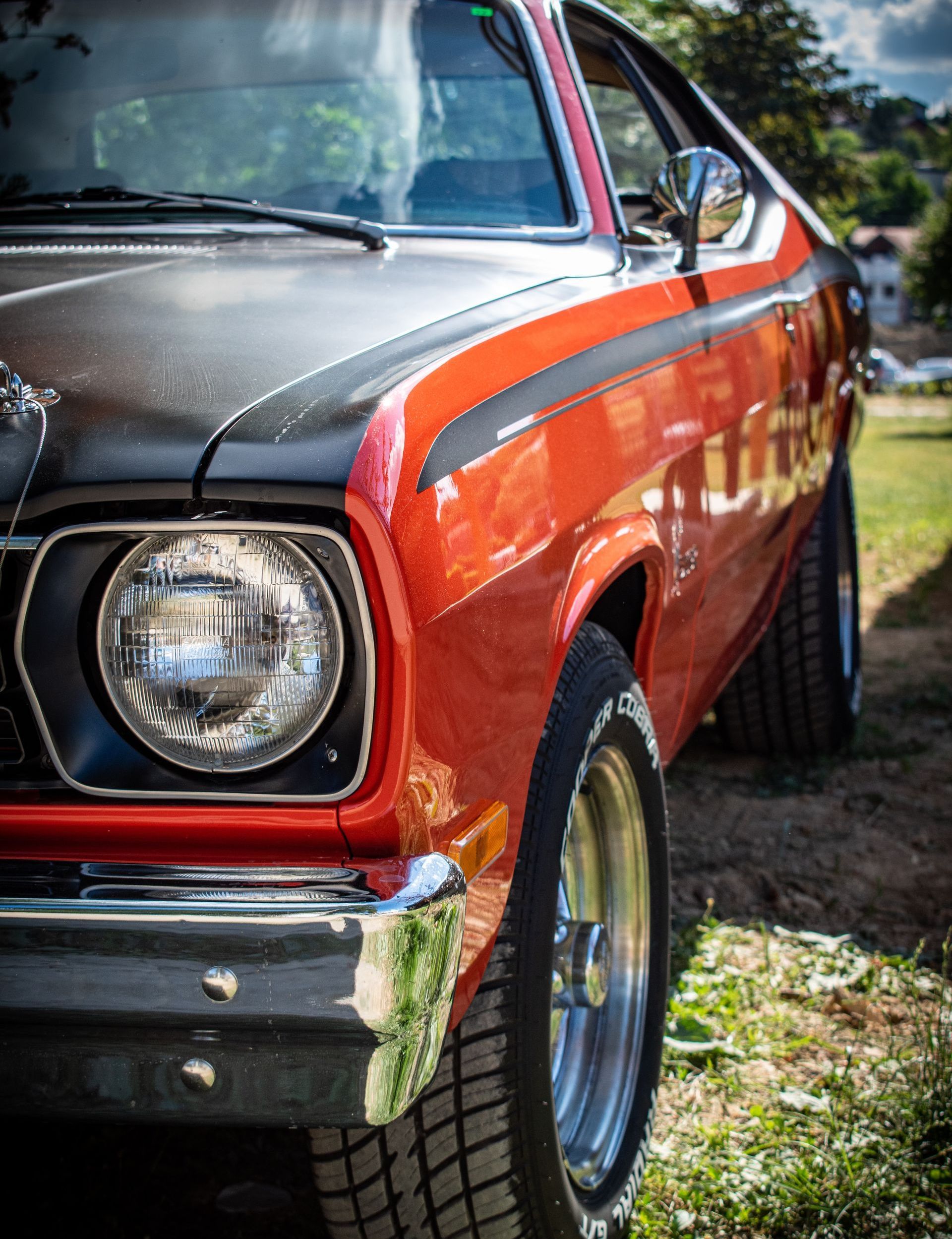 Close-up of a vintage orange and black muscle car, with chrome accents and a black racing stripe, parked outside on a sunny day.