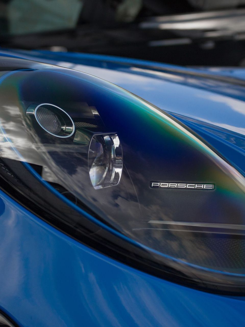 Close-up of a blue car headlight with a Porsche logo, showing a rainbow-like iridescent effect on the lens.