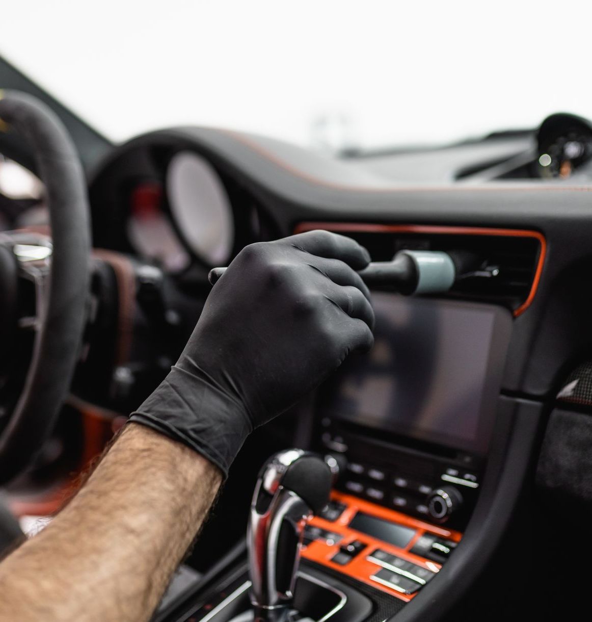 A person wearing a black glove uses a brush to clean a car's dashboard vents; interior shot with orange accents.