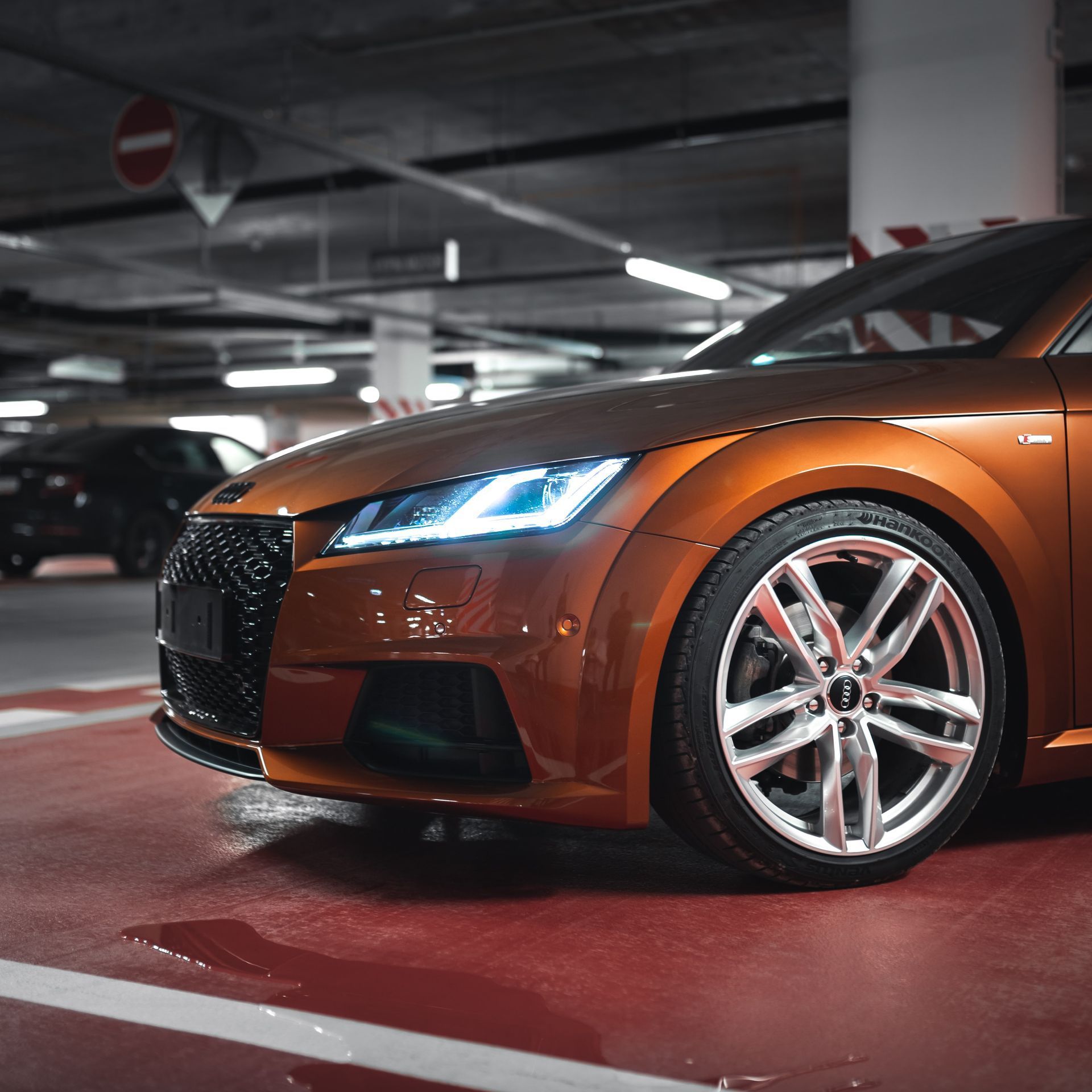Orange Audi TT sports car parked in a dim parking garage, with another car in the background.