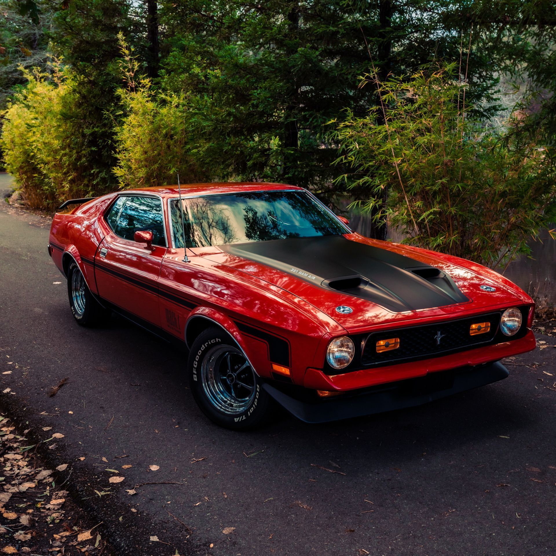 Red 1971 Ford Mustang Mach 1 parked on a paved road with trees in the background, black hood, and black racing stripes.