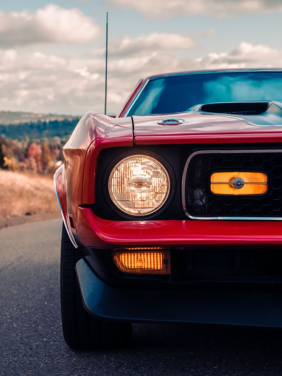 Red classic muscle car's front end with headlight and turn signal lit, parked on a road. Black spoiler and a cloudy sky background.