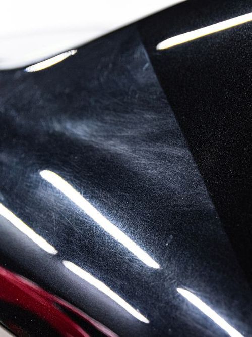 Close-up view of a shiny black car surface showing swirls and scratches, with bright reflections highlighting the imperfections.