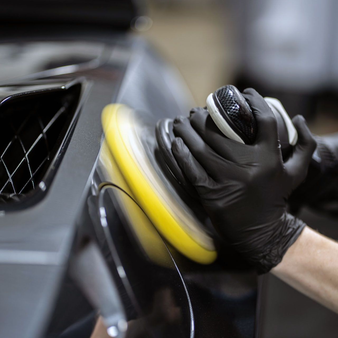 Person wearing black gloves polishing a black car with a rotating yellow buffer.