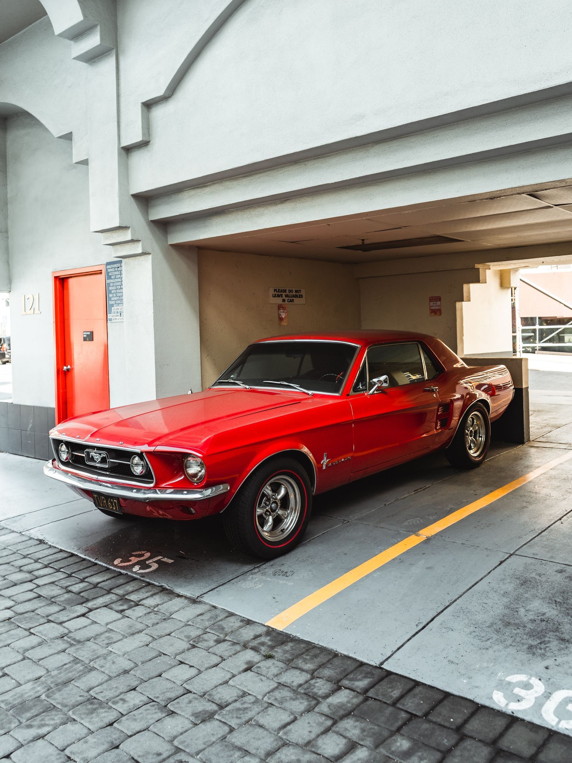 Red classic Ford Mustang parked in a garage. The car is surrounded by gray concrete and a yellow line.