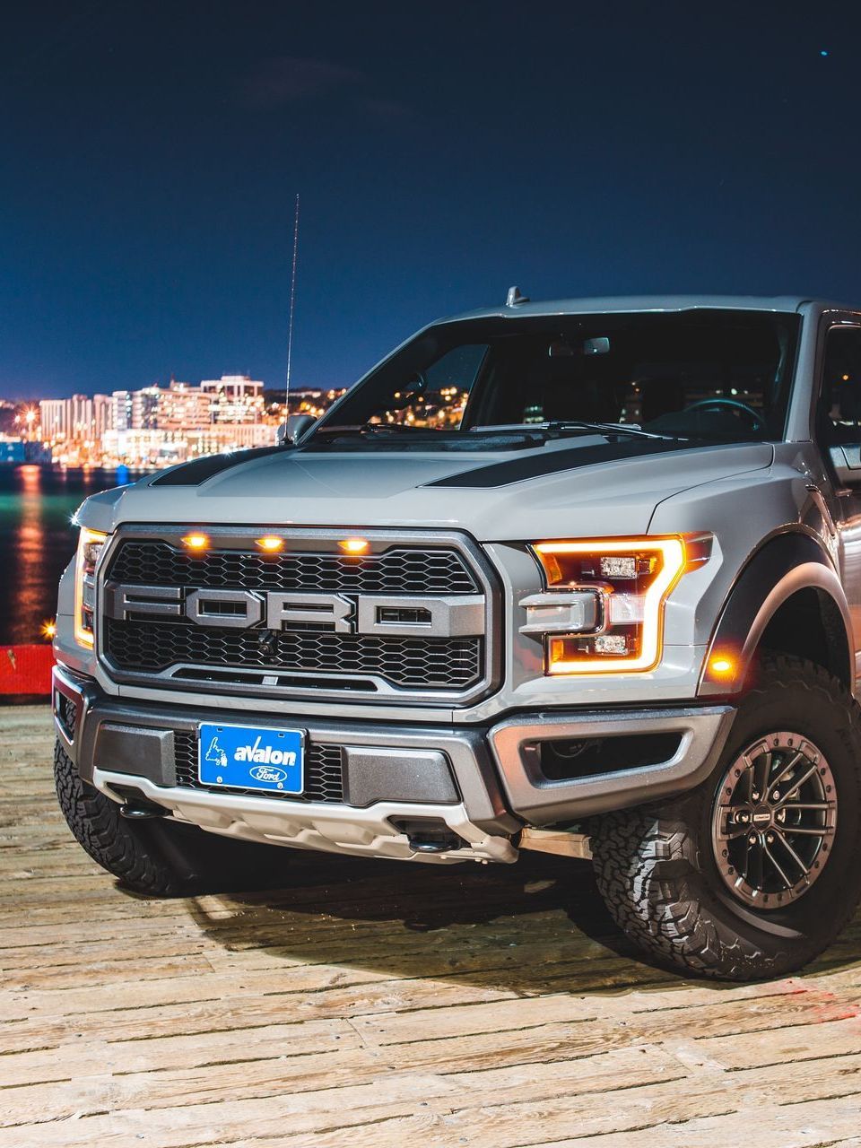 Silver Ford Raptor truck parked, illuminated at night with city lights in the background.