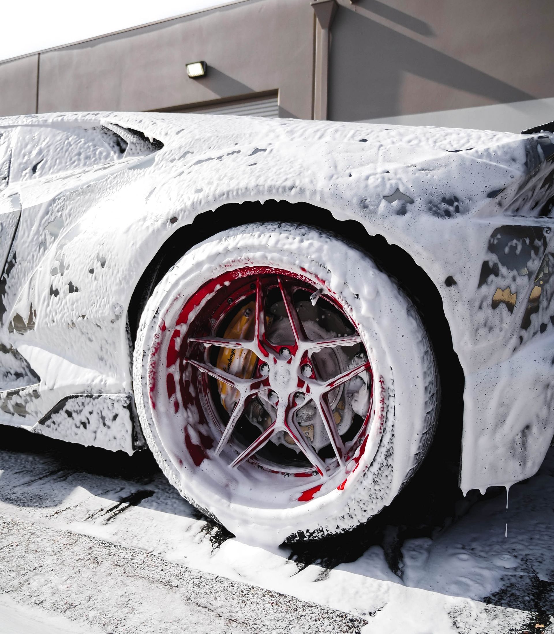 Car covered in white foam with a red and silver wheel. The car is parked outside in front of a building.