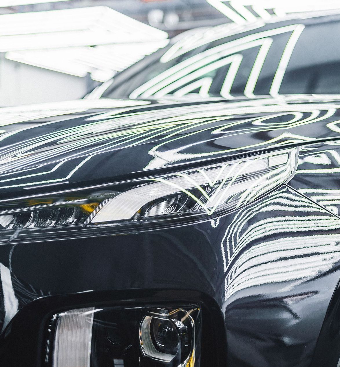 Black car reflecting overhead lights, showcasing headlights and hood details.