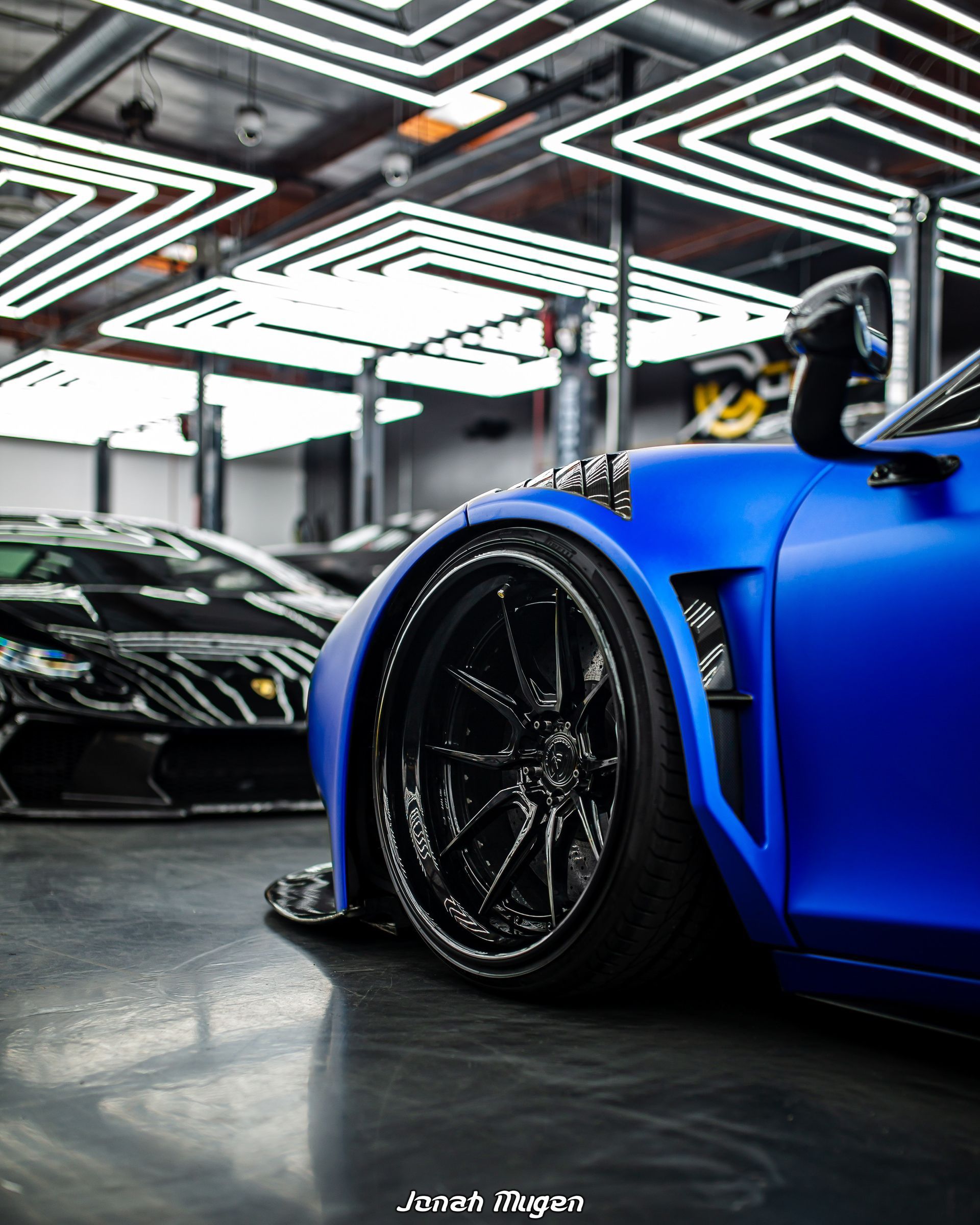 Blue sports car with black wheels, low to the ground, parked inside a bright garage.  Another car is in the background.