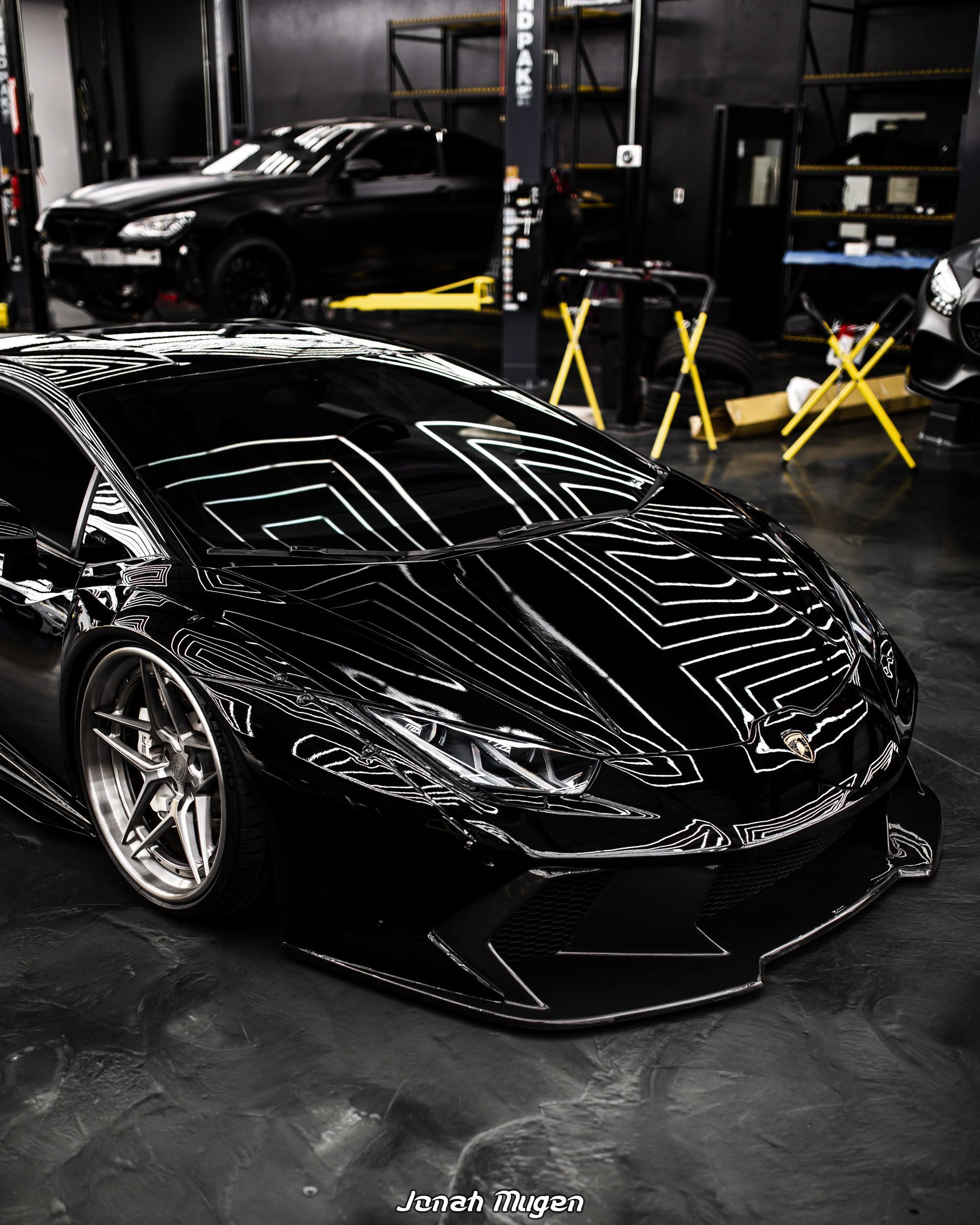 Black Lamborghini sports car in a garage with another vehicle raised on a lift in the background.