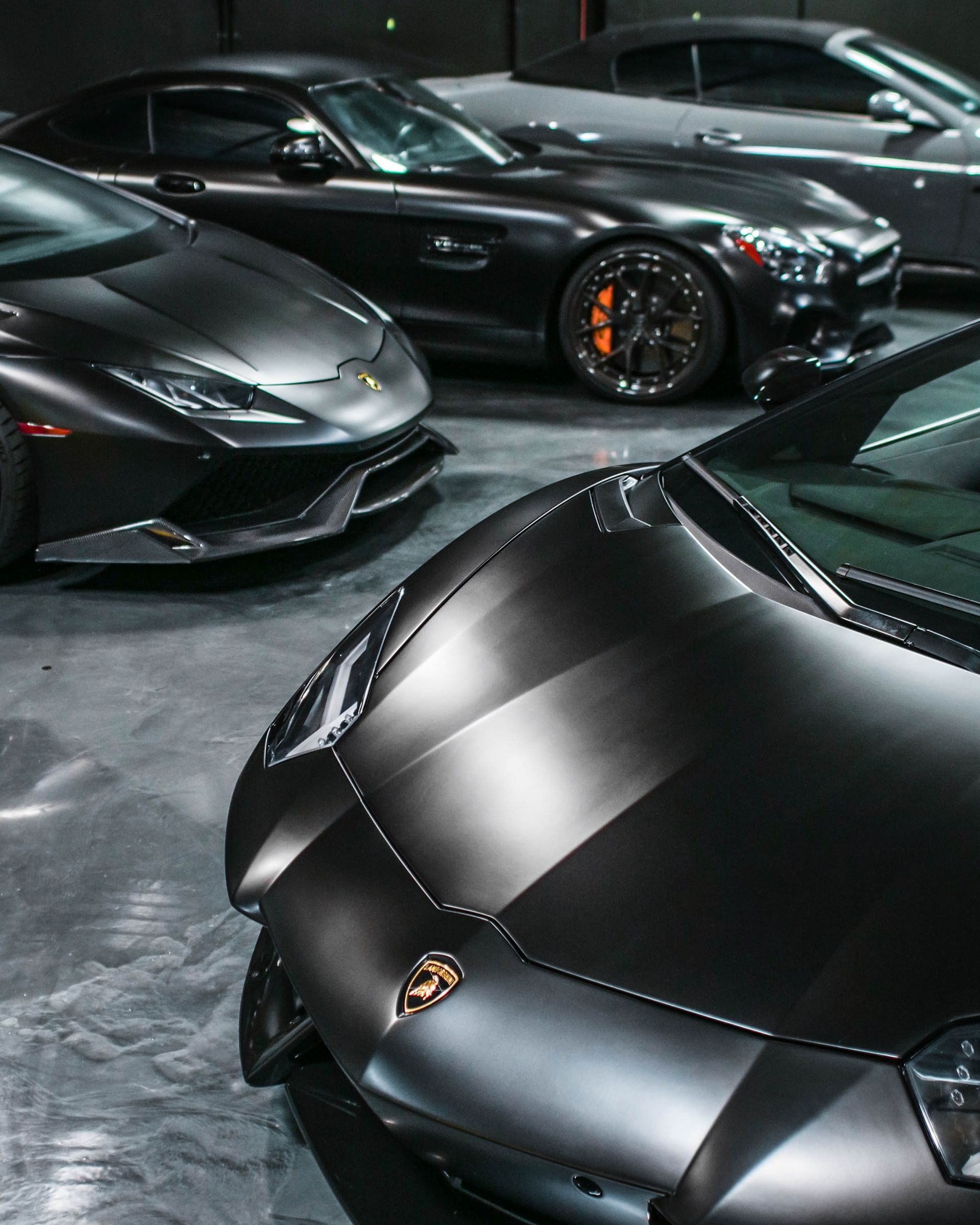 Several luxury black cars parked inside a dimly lit garage, including a Lamborghini, with detailed hoods and rims.