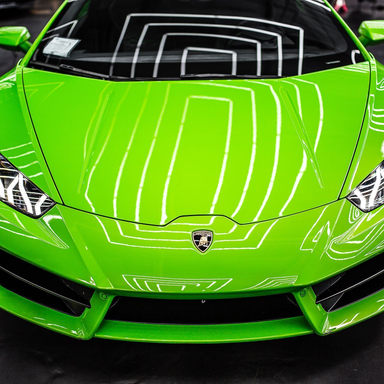 Bright green Lamborghini car, front view, reflecting overhead lights. The car has a prominent hood with a logo.