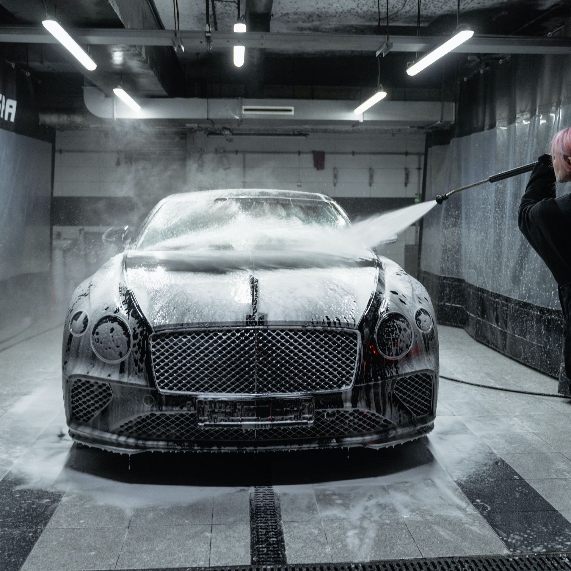 A person sprays a luxury black car, covered in soap, inside a car wash.