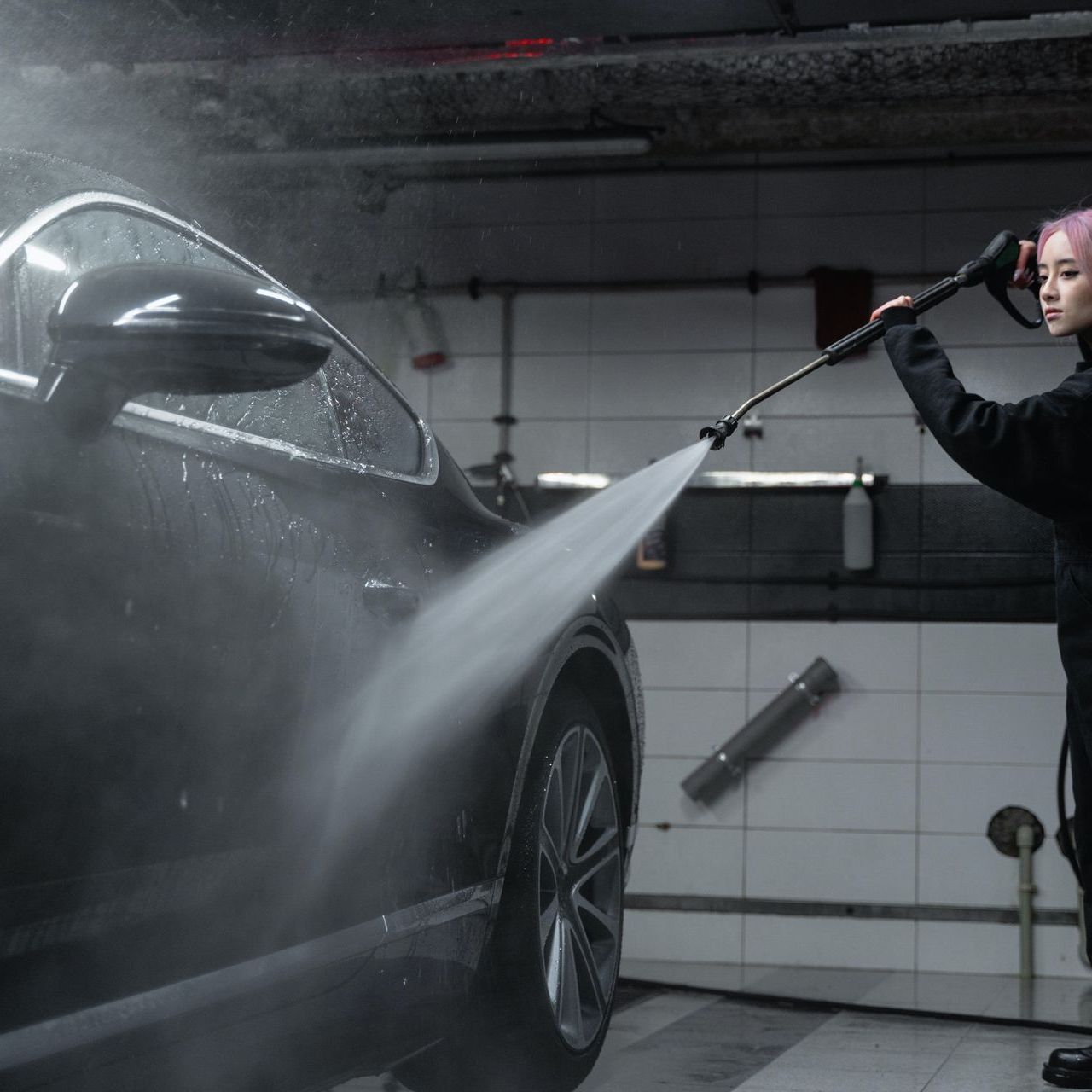Woman with pink hair washes a dark car with a pressure washer in a garage. Water sprays across the car.