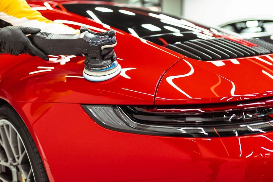 A person polishing a red car with a power polisher in a well-lit setting.