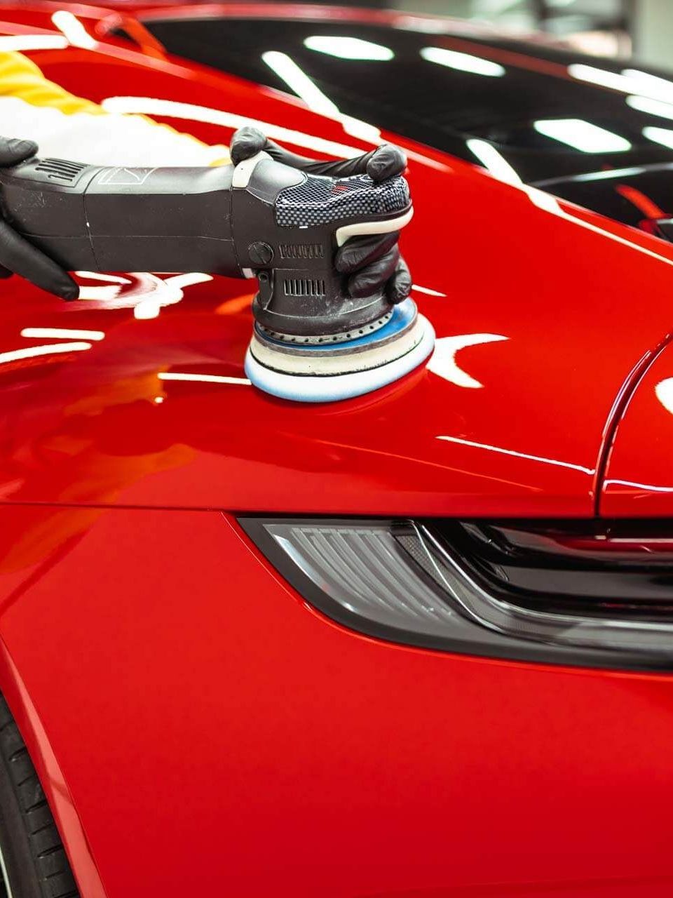 A person polishing a shiny red car with a power buffer. The car's black taillight is visible.