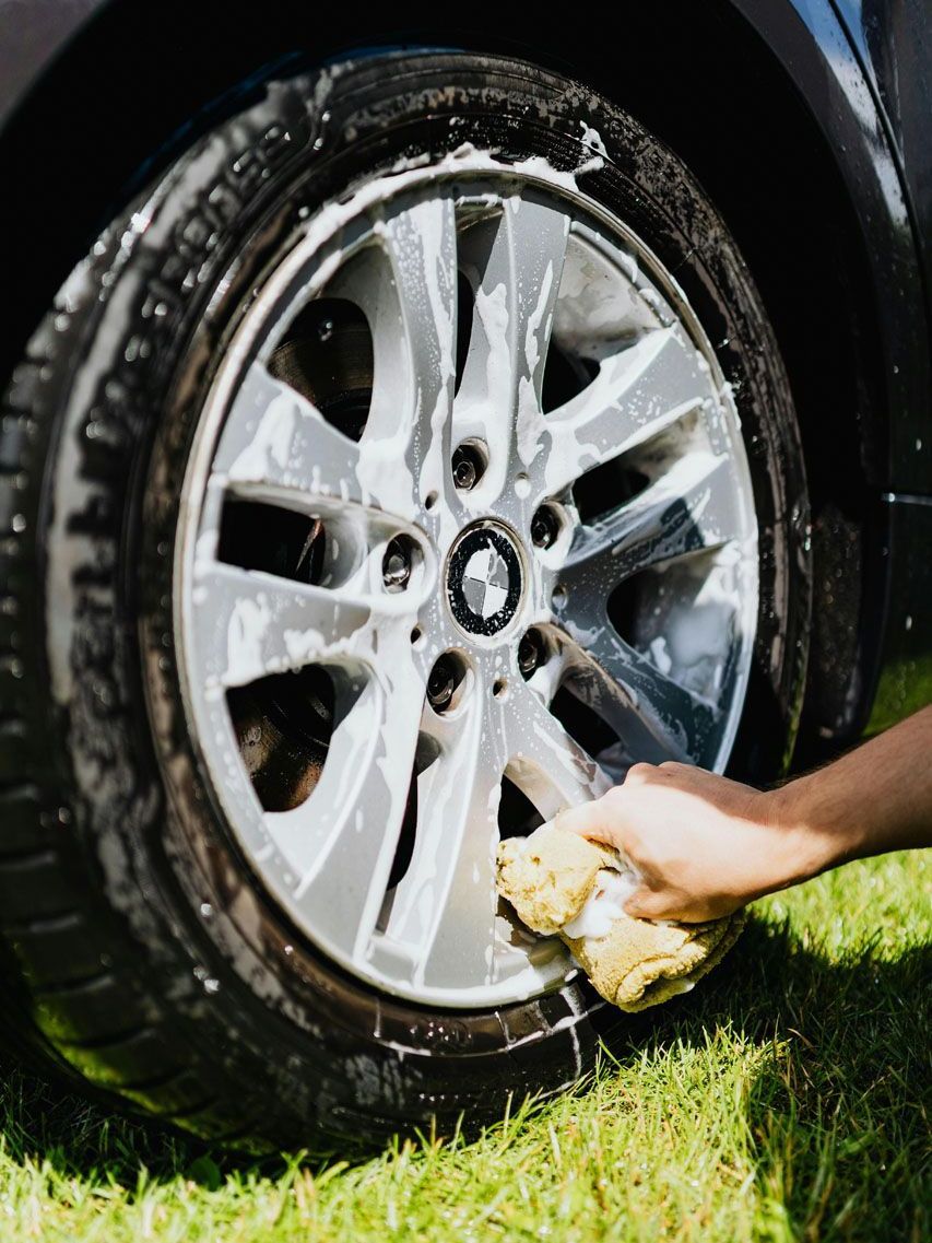 Person washing a car tire and rim with a soapy sponge on green grass.
