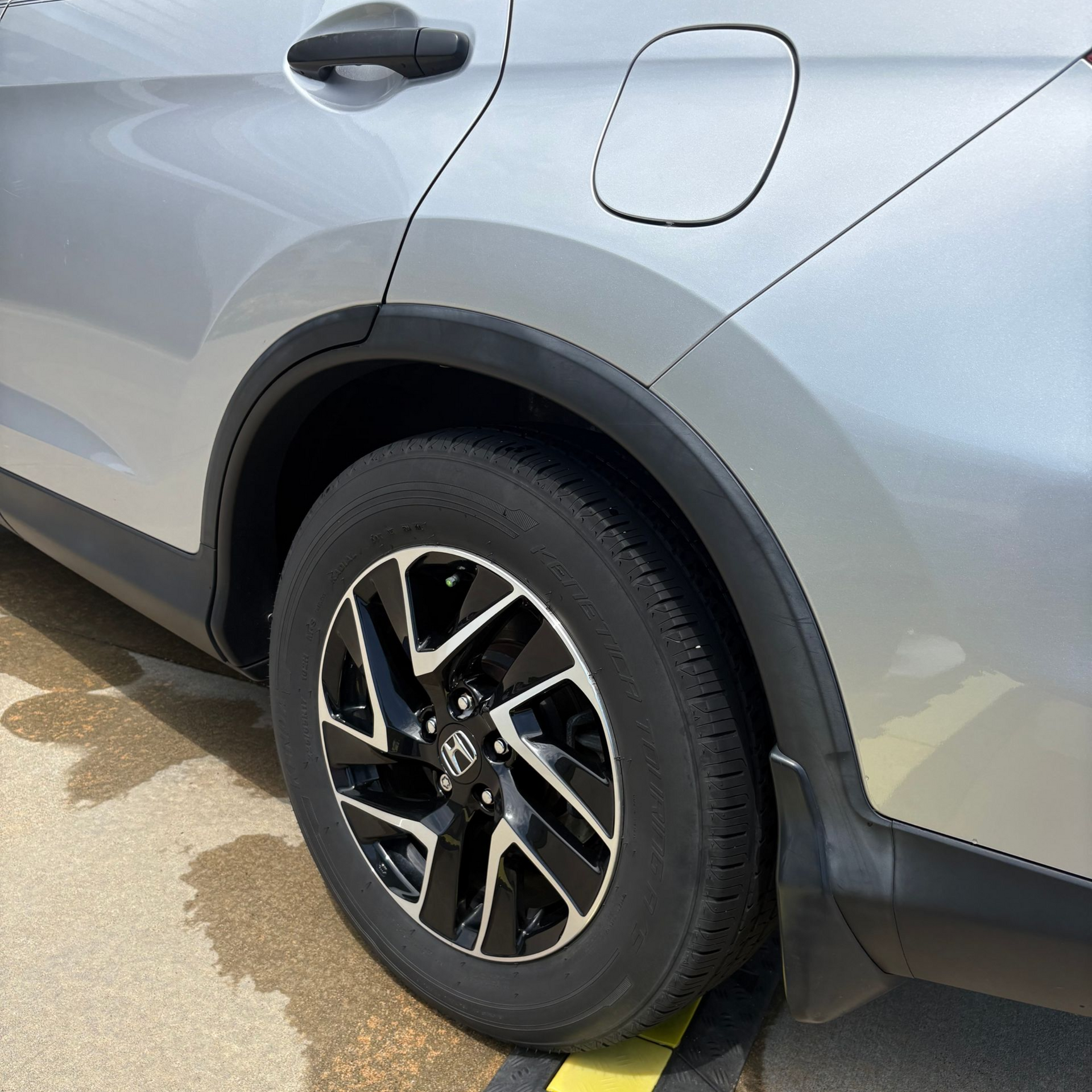 Silver car's rear wheel. Black tire and rim with black wheel well and door handle.