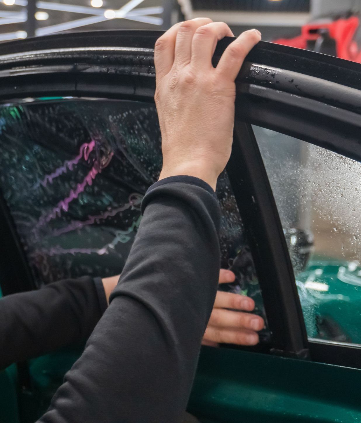 Person's hands applying window tint to a car's side window. Hands are dark against the window film.