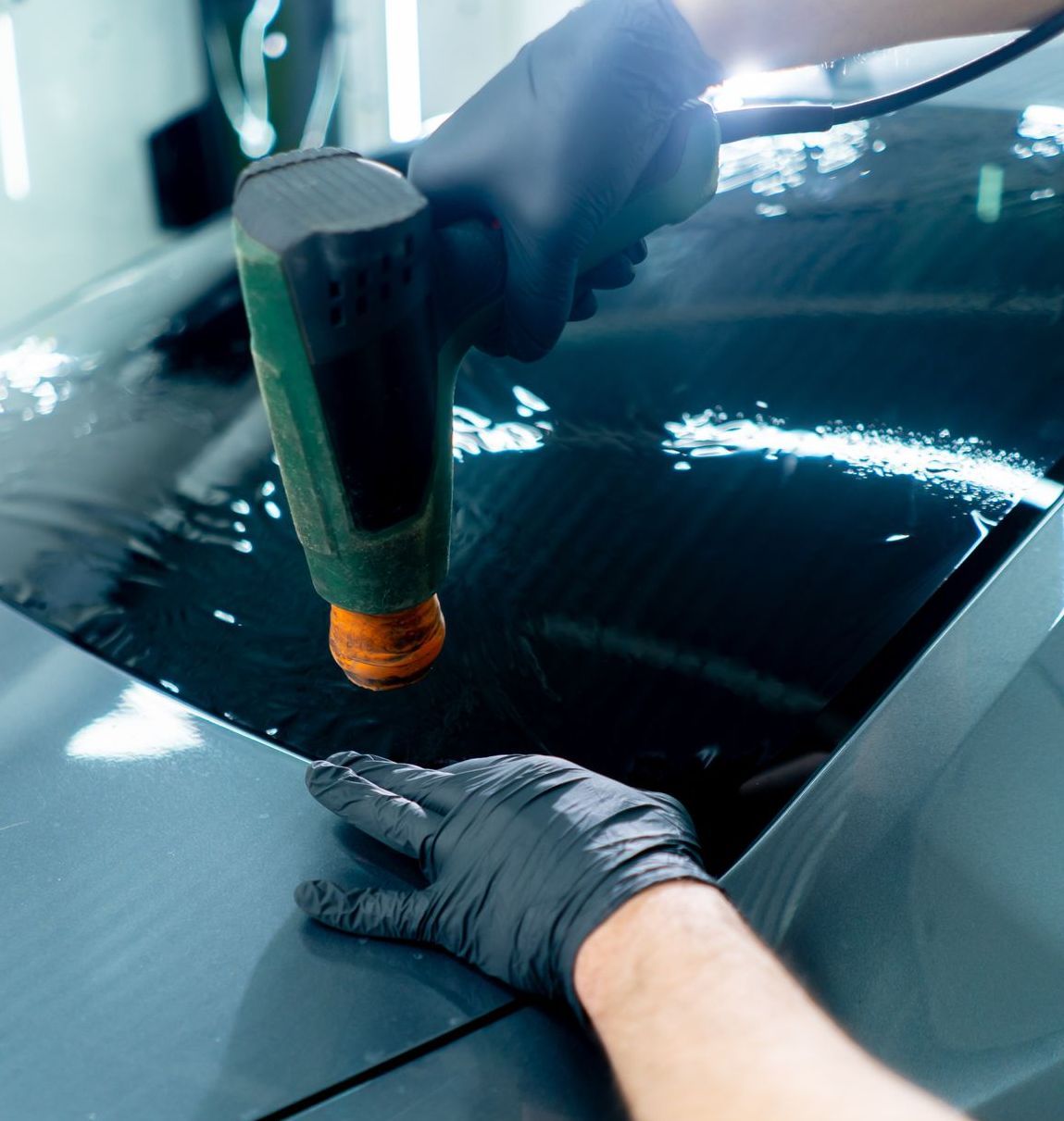 A person applying tint to a car window, using a heat gun and wearing black gloves.