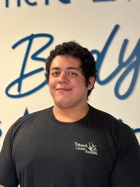 Man with curly hair in a gray t-shirt smiles in front of a blue and white sign.