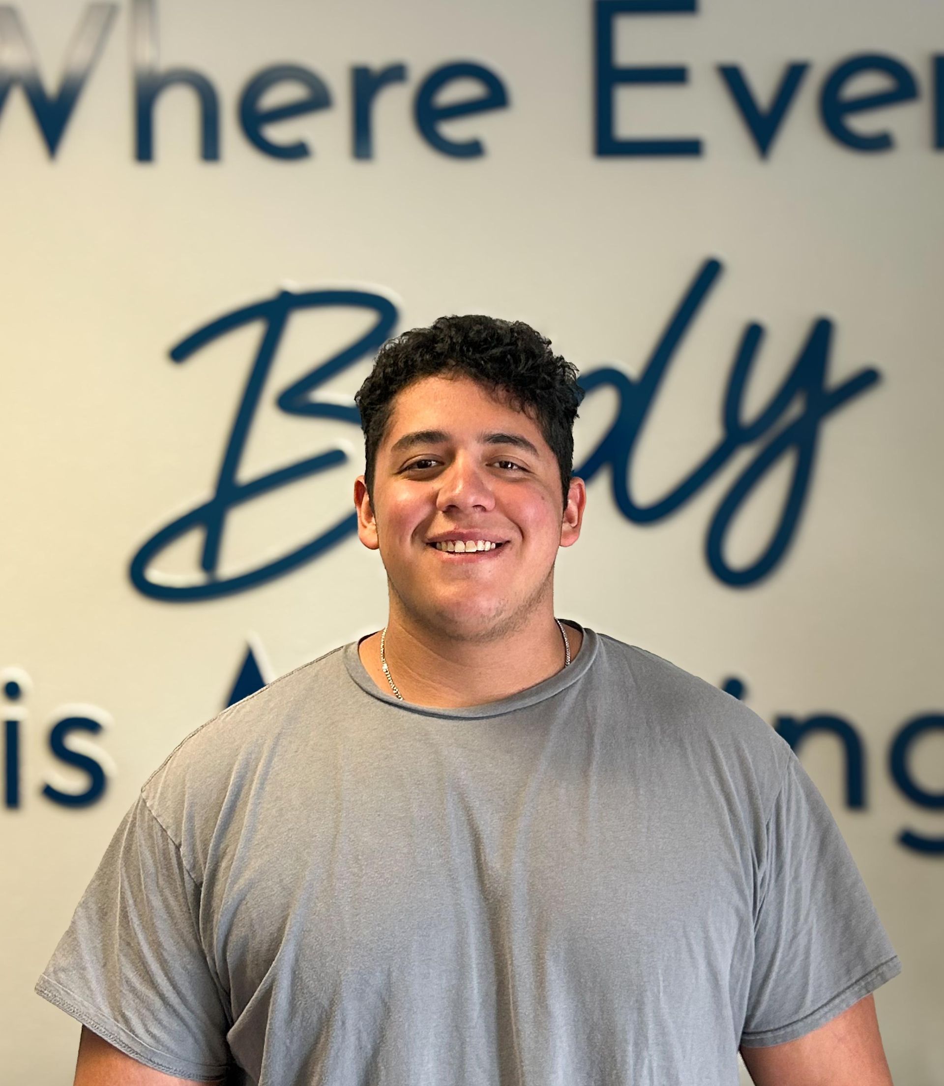 Man with curly hair in a gray t-shirt smiles in front of a blue and white sign.