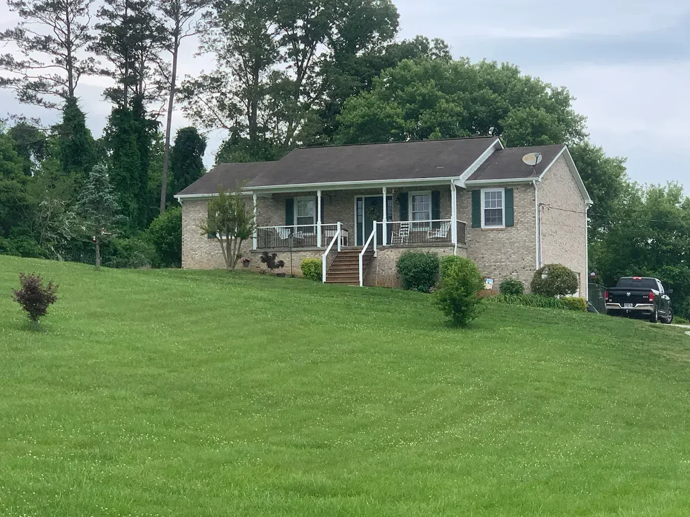 Brick ranch house on a green hill, with a porch, steps, and a car parked nearby.