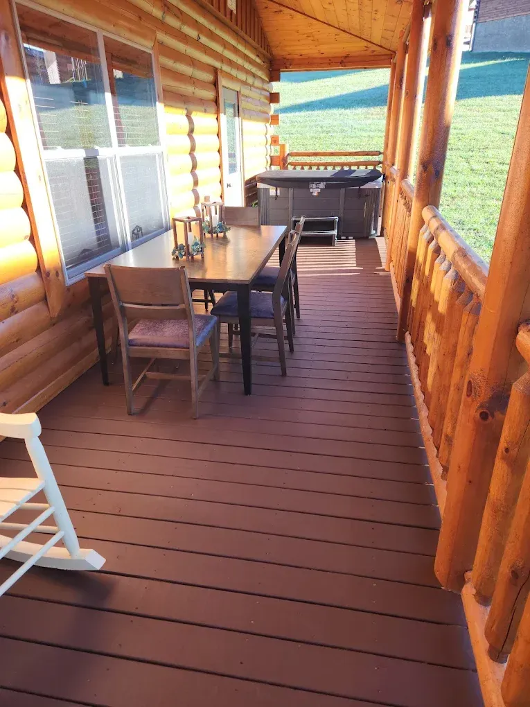 Wooden porch with table and chairs, leading to a hot tub. Brown wood, sunny day.