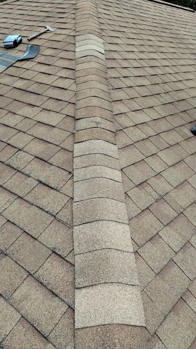 A hammer and roofing materials sit on a tan shingled roof next to a finished ridge cap installation.