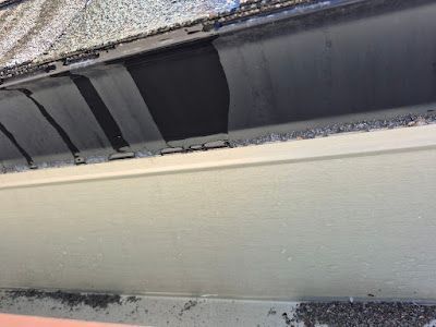 Close-up of a damaged roof edge, showing missing shingles revealing dark underlayment above a beige fascia board.